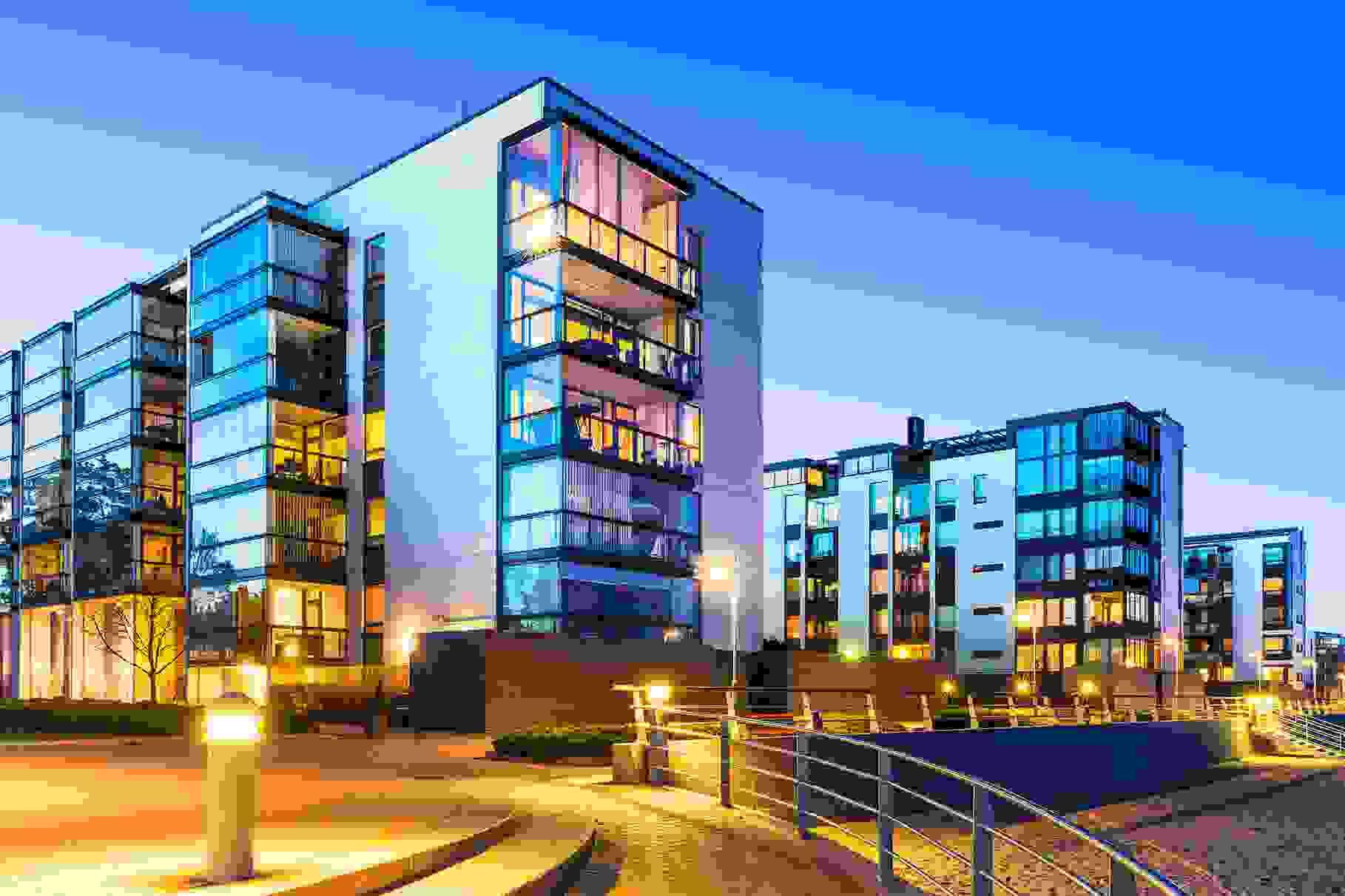 Modern apartment buildings at dusk, lit up by warm-coloured street lights.