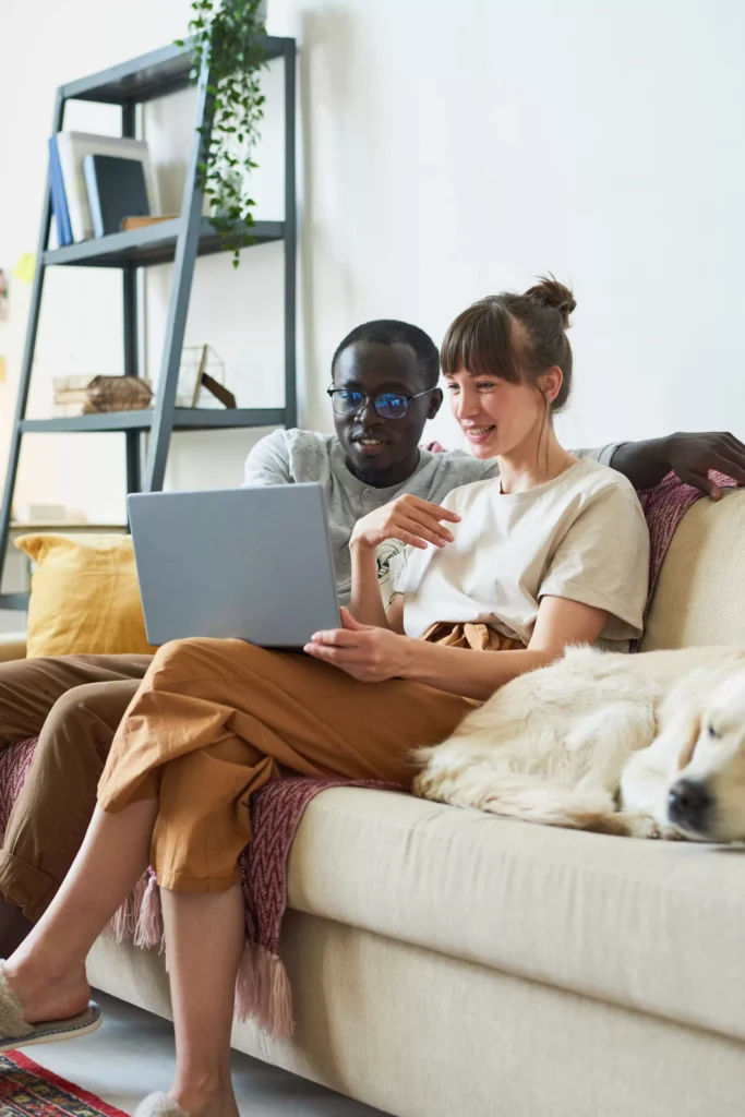 A couple sitting on a sofa looking at a laptop, with a dog lying beside them